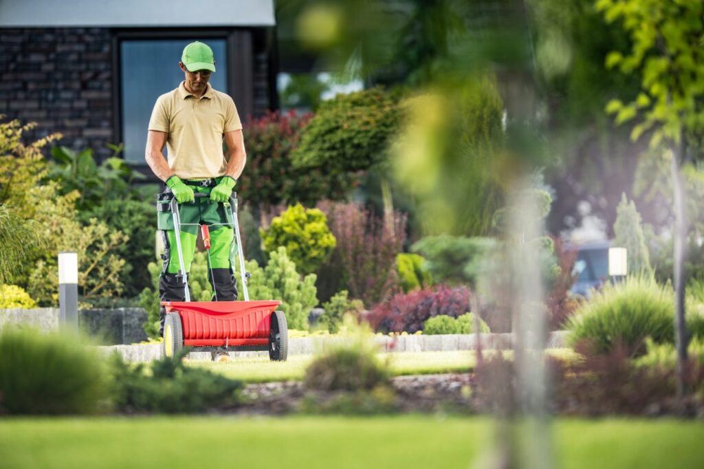 A person in a green cap and gloves is swiftly pushing a red lawn spreader across a well-maintained Lakeland garden. They wear a beige shirt and green pants, with shrubs, a house, and garden lights in the background.