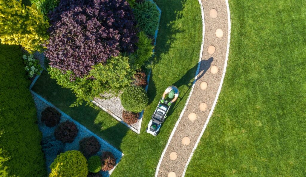 Aerial view of a person providing fast lawn care with a green and black lawnmower in Auburndale. The garden is landscaped with shrubs, a curved gravel path adorned with round stepping stones, and lush greenery.