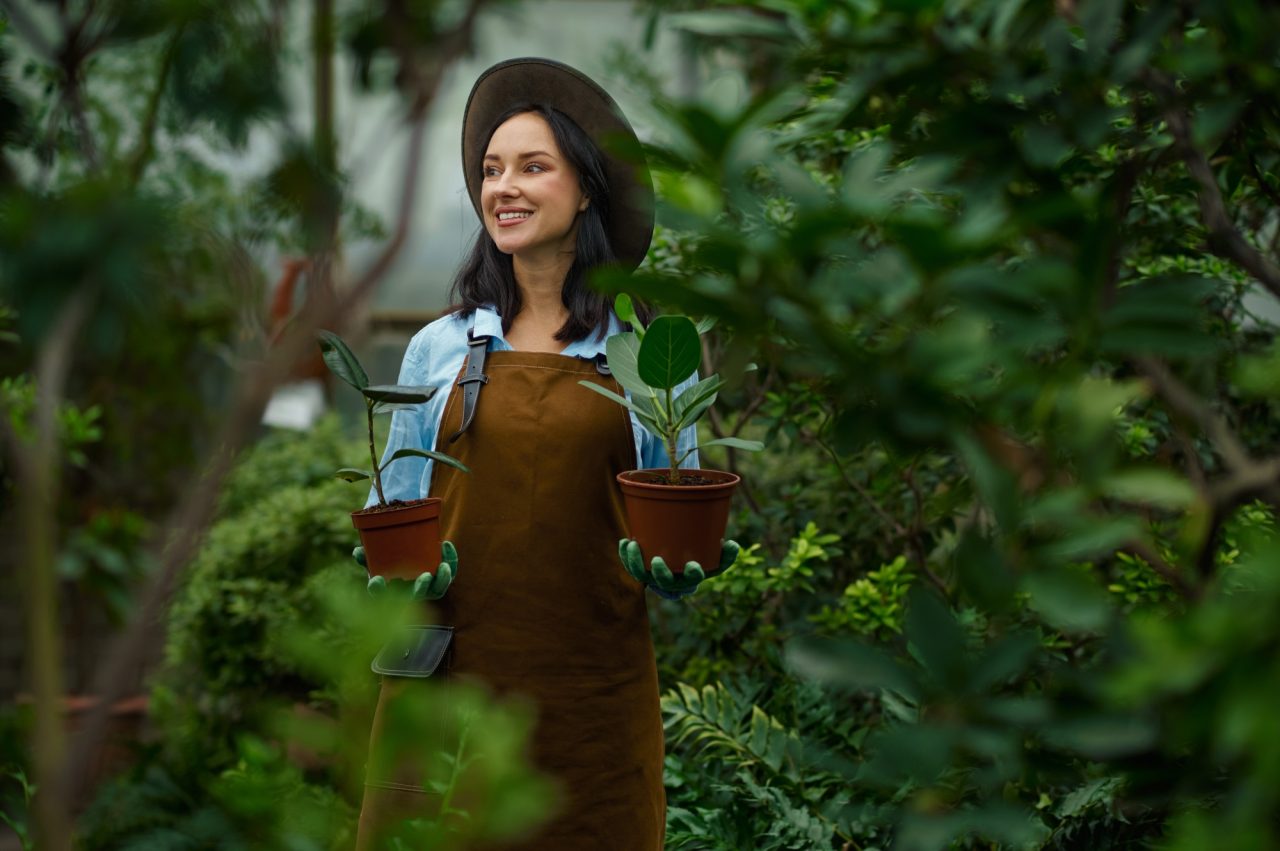 A person wearing a brown apron and a hat smiles while holding two potted plants in a lush, green garden in Winter Haven. Surrounded by various types of foliage, they exude joy as if they've mastered the art of fast lawn care.